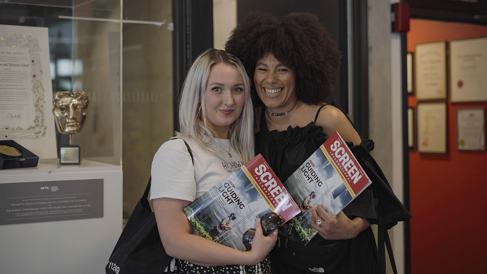 Two women posing for camera next to NFTS Queens Award cabinet